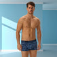 En lyshudet mand med kort brunt hår står i et blåt rum iført Henderson-boxershorts i marineblåt mix med camouflagemønstre og lavet af blød bomuld, vendt fremad med et neutralt udtryk midt i lyse geometriske vægmønstre.