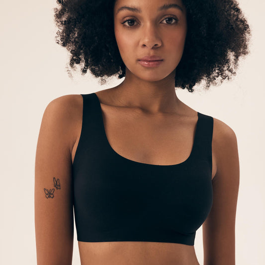 En kvinde med krøllet hår bærer en sort, sømløs, fornemmelsefyldt Henderson sports-bralette med udtageligt vat. Hun har et neutralt udtryk og en lille sommerfugletatovering på sin venstre overarm, sat op mod en almindelig lys baggrund.