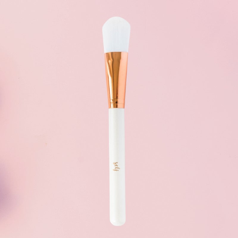 En flad, vegansk Natural Self-børste med hvide børstehår, en metallisk hylster i rosaguld og et hvidt skaft med påskriften "self" er centreret på en lyserød baggrund - ideel som ansigtsmaske redskab eller Facial Treatment Brush.