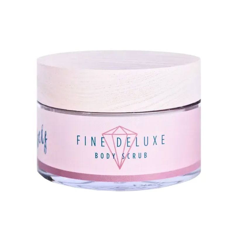 En klar, rund krukke fra Natural Self med et let træstruktureret låg indeholder bodyscrub med dødehavssalt. Den lyserøde etiket har en rød diamantgrafik og en blågrøn tekst, hvor der står FINE DELUXE BODY SCRUB. Baggrunden er helt hvid.