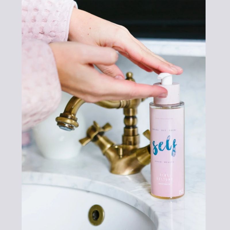 En person i lyserød morgenkåbe doserer Natural Self body wash fra en lyserød pumpeflaske over en hvid marmorvask med en messinghane, mens hænderne er klar til at fange produktet - perfekt til følsom hud på denne stilfulde badeværelsesbordplade.
