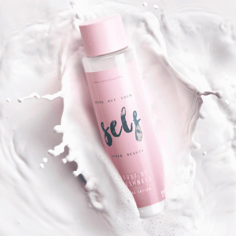 En pink cylinderformet flaske Natural Self med påskriften "self" og "inner beauty" hviler diagonalt på en hvid overflade blandt hvirvlende hvide væskesprøjt, der leder tankerne hen på frisk, blød hud. Den pastelfarvede hætte fremhæver mærkets minimale og elegante design.