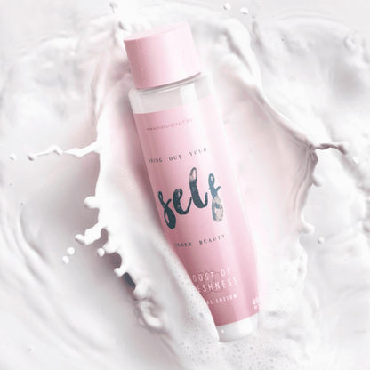 En pink cylinderformet flaske Natural Self med påskriften "self" og "inner beauty" hviler diagonalt på en hvid overflade blandt hvirvlende hvide væskesprøjt, der leder tankerne hen på frisk, blød hud. Den pastelfarvede hætte fremhæver mærkets minimale og elegante design.