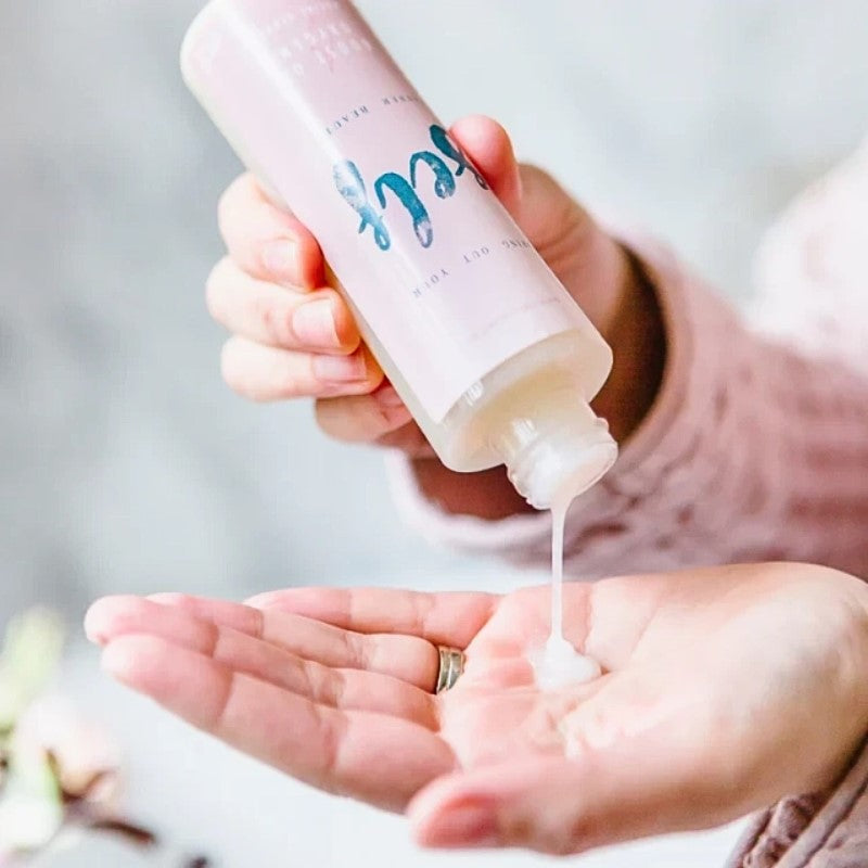 En hånd hælder cremet hvid ansigtsscrub fra en pink Natural Self-flaske med blågrøn tekst, der antyder naturlige ingredienser, i en andens håndflade. Personen bærer et lyserødt strikærme og en ring; baggrunden er let sløret.