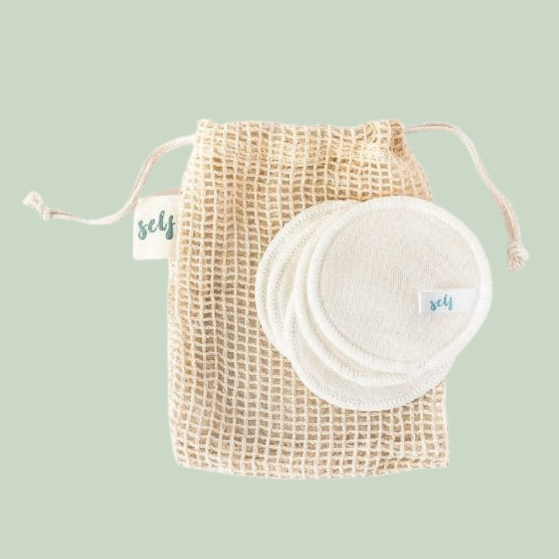 En beige netpose med snoretræk mærket "Natural Self" indeholder flere økologiske bambus genanvendelige vatrondeller med små "Natural Self"-mærker, der er arrangeret pænt på en lysegrøn baggrund.