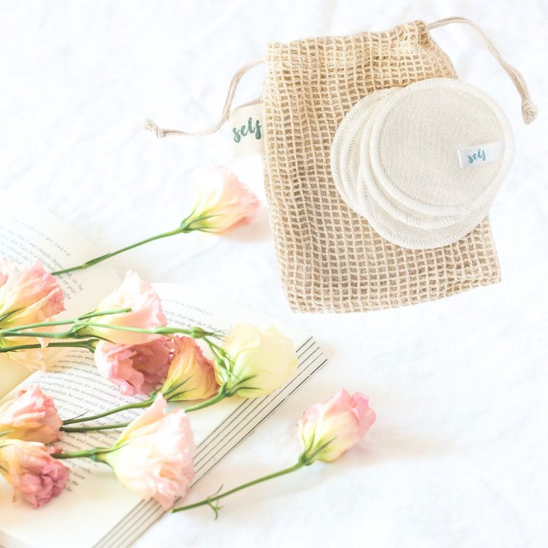 En blød netpose med snor rummer flere genanvendelige vatrondeller fra Natural Self, med et lille "self"-mærke synligt. Lyserøde blomster hviler på en åben bog og skaber en frisk, miljøvenlig scene - ideel til sensitiv hud.