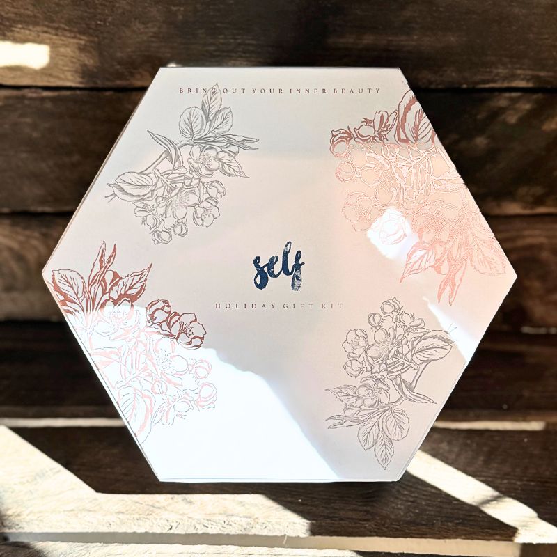En sekskantet æske fra Natural Self med blomsterillustrationer, blå "self"-bogstaver og "BRING OUT YOUR INNER BEAUTY" plus "HOLIDAY GIFT KIT" på låget; denne gavesæt hudpleje hviler på træ i blidt sollys.