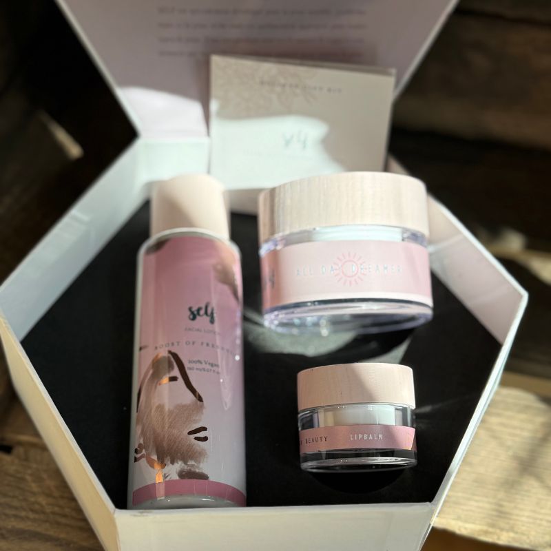 En hvid, sekskantet gaveæske fra Natural Self indeholder tre veganske hudplejeprodukter i rosa og hvid - en høj flaske, en mellemstor krukke og en lille "LIPBALM"-krukke - udstillet på sort underlag med sollys på sættet og et kort i låget.