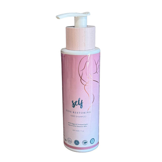 En cylindrisk, pink og minimalistisk flaske med Natural Self Self Shine Restoring Hair Shampoo med en pumpedispenser. Etiketten fremhæver den veganske formel og de naturlige ingredienser med certificeringsikoner og detaljer i bunden.