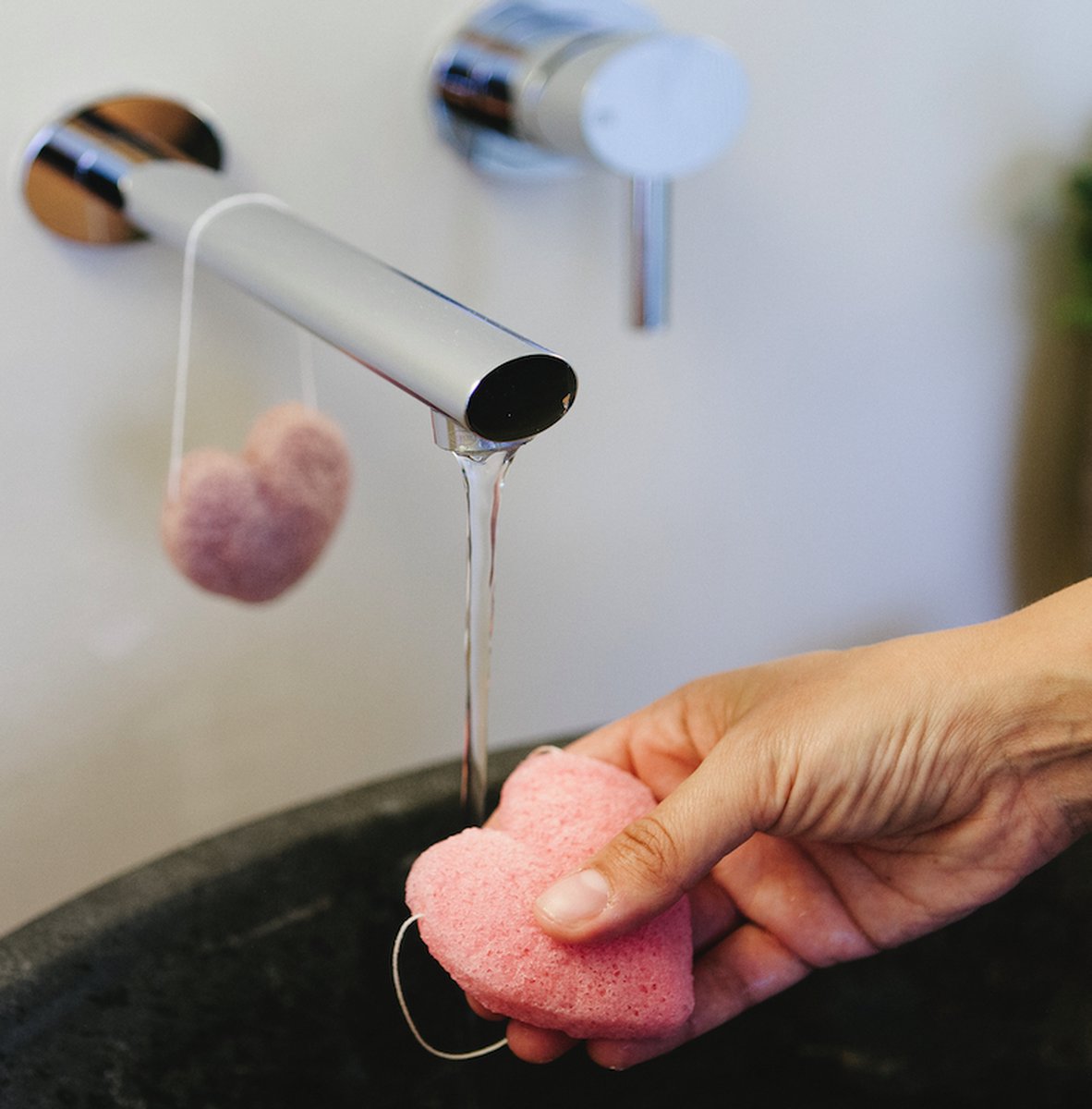 En hånd holder en lyserød, hjerteformet Natural Self konjac svamp under en rindende vandhane over en mørk vask. I baggrunden hænger en anden lignende svamp lavet af naturligt konjac-pulver til ansigtsrens i en snor fra vandhanehåndtaget.