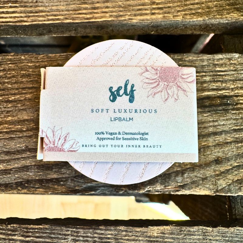 En rektangulær Natural Self lipbalm-pakke hviler på forvitret træ. På etiketten står der: "Soft Luxurious Lipbalm" med blomstermotiver, og der står, at det er en vegansk læbepomade, godkendt af hudlæger og velegnet til følsom hud.