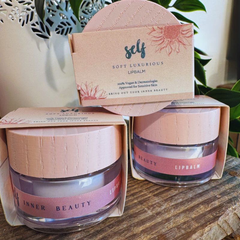 To lyserøde krukker med etiketten Lipbalm INNER BEAUTY fra Natural Self står side om side. Over dem står der "self SOFT LUXURIOUS LIPBALM" på en matchende pink æske, som fremhæver, at denne veganske læbepomade er velegnet til følsom hud.