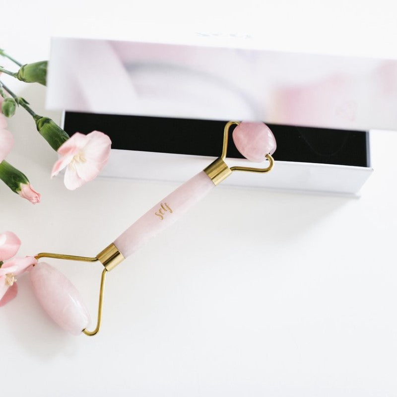 En Natural Self Rose Quartz Facial Roller med gulddetaljer ligger delvist inde i en hvid æske. Lyserøde blomster til venstre giver et delikat touch, mens ansigtsmassagerullen har store og små sten til mindske hævelser.