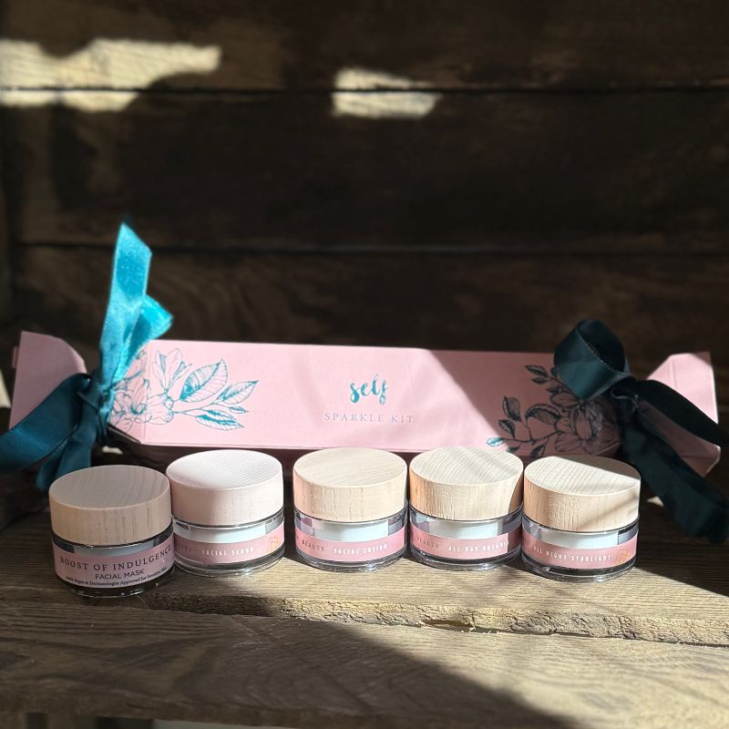 Fem små Natural Self-krukker med lyse trælåg, mærket til ansigtspleje, er stillet op foran en pink blomstret Self Sparkle Kit gavesæt hudpleje-æske bundet med blå bånd mod en rustik træbaggrund.