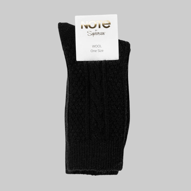Et par Note sorte uldstrømper, pænt foldet med en hvid etiket med teksten NOTE Superior i guld og WOOL One Size i gråt, vist på en almindelig lysegrå baggrund.