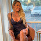 En smilende blond kvinde med synlige tatoveringer iført en gennemsigtig sort lingerikjole fra Obsessive sidder på en brun stol ved et stort vindue. Det ene ben er krydset over det andet, og man kan se grønne områder og en parkeret bil udenfor.