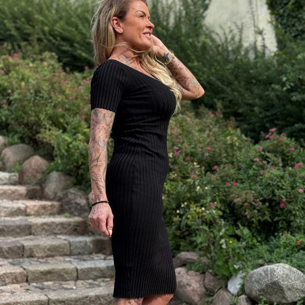 En kvinde med langt blondt hår og synlige tatoveringer smiler, mens hun rører ved sit hår, stående udendørs på en stensti i en rib-strikket midi-kjole med V-udskæring fra Soy & Co, omgivet af grønne buske og lyserøde blomster.
