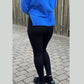 En person står udendørs på en stenbelægning med ryggen til kameraet iført en lyseblå sweater og Soy & Co Sevilla-leggings. Sorte sko, et træhegn og spredte blade er også synlige.