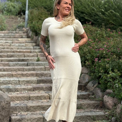 En kvinde med langt blondt hår og tatoveringer på armen står på en stentrappe udendørs og smiler i en tætsiddende, kortærmet MOCHY beige ribstrik kjole. Den ankellange kjole flatterer hendes figur blandt grønne buske og blomster i baggrunden.