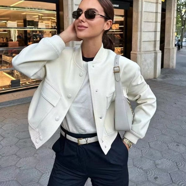 En kvinde står udendørs med hænderne i siden og smiler, klædt i MW Studio solbriller, en tidløst valgt hvid bomberjakke, hvid skjorte, sorte bukser og en hvid håndtaske – perfekt til året mod en urban baggrund.