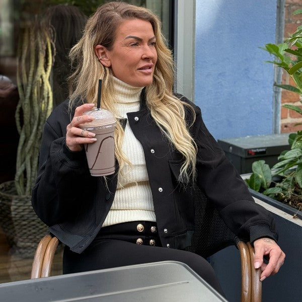 En kvinde med langt bølget blondt hår sidder udendørs og smiler, mens hun nyder en chokoladedrik. Hun er iført MW Studio let overtøj - en slags jakke over en hvid rullekrave og sorte bukser med guldknapper. Bag hende er der grønne planter og et vindue.