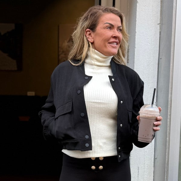 En kvinde med bølget blondt hår står ved en døråbning og smiler til siden. Hun er iført en kort MW Studio-jakke over en hvid rullekrave, sorte bukser med guldknapper og holder en chokolademilkshake i sin højre hånd.