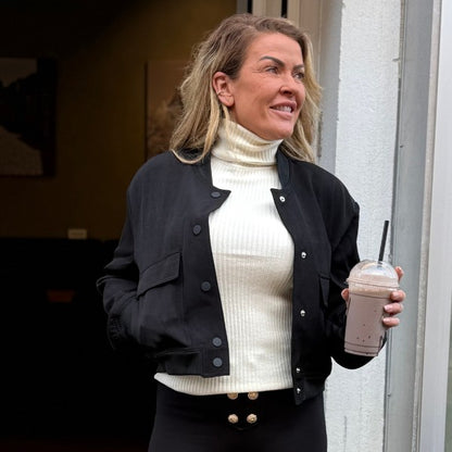 En kvinde med bølget blondt hår står ved en døråbning og smiler til siden. Hun er iført en kort MW Studio-jakke over en hvid rullekrave, sorte bukser med guldknapper og holder en chokolademilkshake i sin højre hånd.