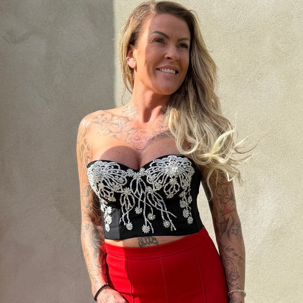 En smilende kvinde med langt, bølget blondt hår står op ad en almindelig væg iført en Soy & Co stropløs sort bustier med hvidt broderi og en højtaljet rød nederdel. Sollyset kaster skygger bag hende og fremhæver hendes tatoverede arme og bryst.