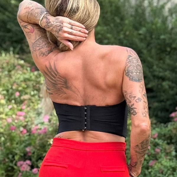 En kvinde med tatoverede arme og ryg, der ser væk og rører ved sit hoved, bærer en Soy & Co stropløs bustier med kroge i ryggen og knaldrøde bukser. Grønt løv og lyserøde blomster sløres i baggrunden.