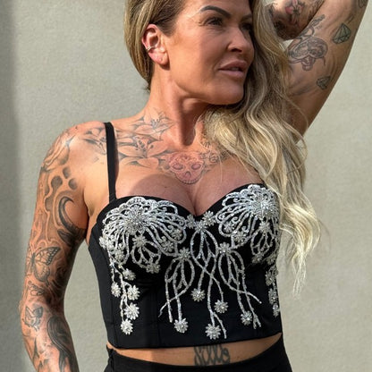 En kvinde med langt blondt hår og tatoveringer poserer mod en almindelig baggrund iført en sort bustier-top fra Soy & Co dekoreret med sølvperler og håndlavet blomsterdesign, hendes højre arm er løftet bag hovedet, mens hun kigger til højre.