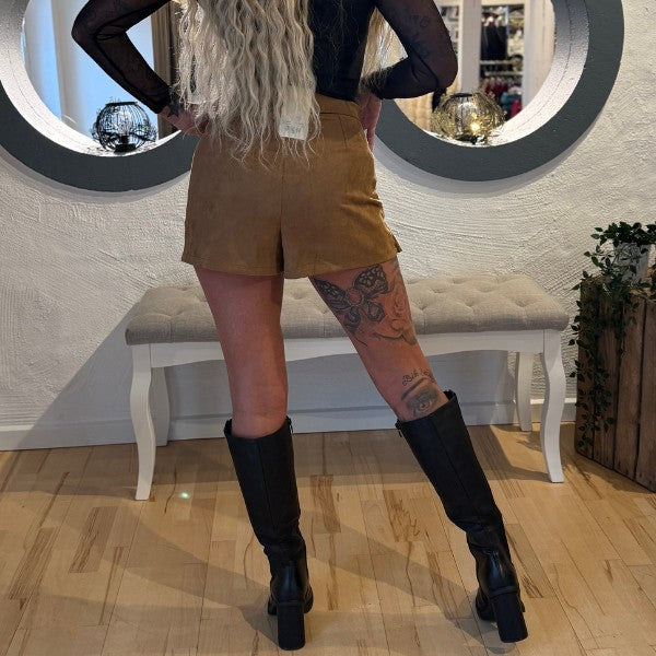 En kvinde med langt, bølget, blond hår står indendørs og vender væk. Hun har en kobberrød, gennemsigtig, sort langærmet top på, kamelfarvede højtaljede ruskinds shorts og sorte knæhøje støvler. Tatoveringer pryder hendes venstre ben nær en bænk og runde spejle på en hvid væg.
