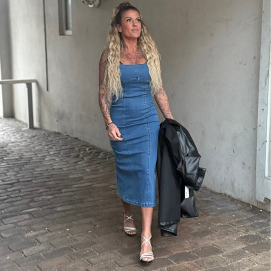 En kvinde med langt, bølget blond hår går forbi en hvid væg i en Girl Vivi denimkjole, der fremhæver hendes tidløse figur og skaber en flatterende effekt. Hun smiler let, bærer en sort jakke og hvide sandaler med remme.