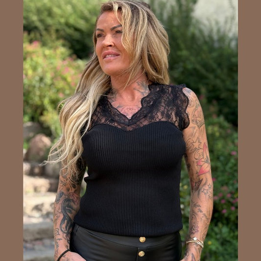 En tatoveret kvinde med langt blondt hår smiler udendørs iført en J&H Fashion-top med ribkant og sorte bukser med guldknapper. Baggrunden består af grønne områder og slørede trin.