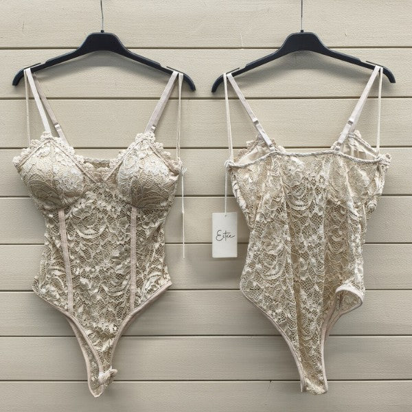 To beige ESTEE-undertøjsbodyer med justerbare stropper hænger på sorte bøjler mod en lys trævæg - den ene viser den formstøbte skål foran, den anden ryggen med et ESTEE-mærke. Begge har en figursyet pasform og er forsynet med indviklede blondemønstre.
