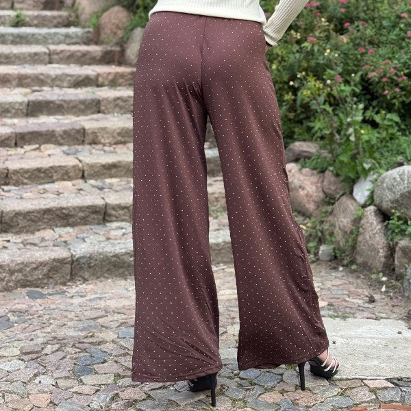 En person står udendørs på en brostensbelagt sti med stentrapper bagved iført Soy & Co brun bukser med små hvide prikker, en cremefarvet langærmet top og sorte sandaler med hæl. Grønne buske og sten omkranser stien.