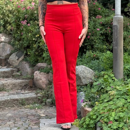 En tatoveret person står udendørs på en stensti iført MOCHY højtaljede, udspilede knaldrøde bukser og sorte sko med åben tå. Frodige grønne buske med lyserøde blomster og klipper er i baggrunden; deres overkrop er beskåret.