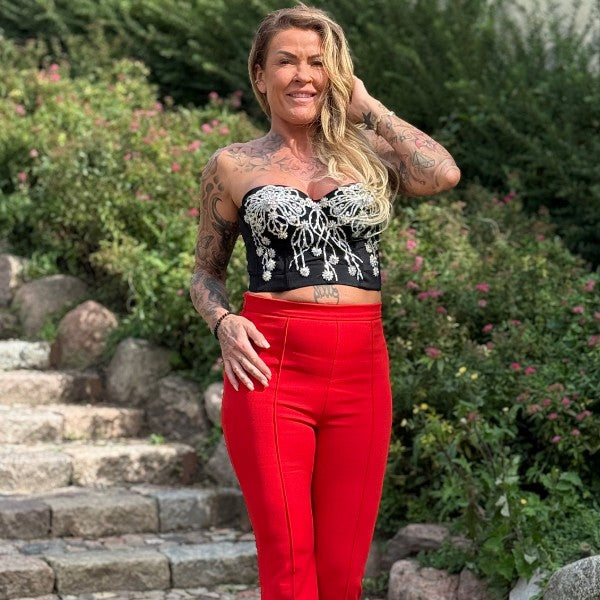 En smilende kvinde med langt blondt hår poserer på en stentrappe iført MOCHYs lyse røde bukser og en sort stropløs top med sirlige hvide broderier. Hendes tatoverede arme og bryst skiller sig ud blandt grønne planter og lyserøde blomster.