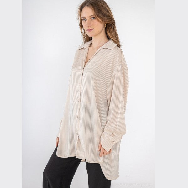 En kvinde poserer på en hvid baggrund iført en løs, lodretstribet lang skjorte fra J&H Fashion i lys beige med sorte bukser. Hun smiler forsigtigt med hånden i lommen og står med en afslappet holdning.