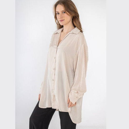 En kvinde poserer på en hvid baggrund iført en løs, lodretstribet lang skjorte fra J&H Fashion i lys beige med sorte bukser. Hun smiler forsigtigt med hånden i lommen og står med en afslappet holdning.