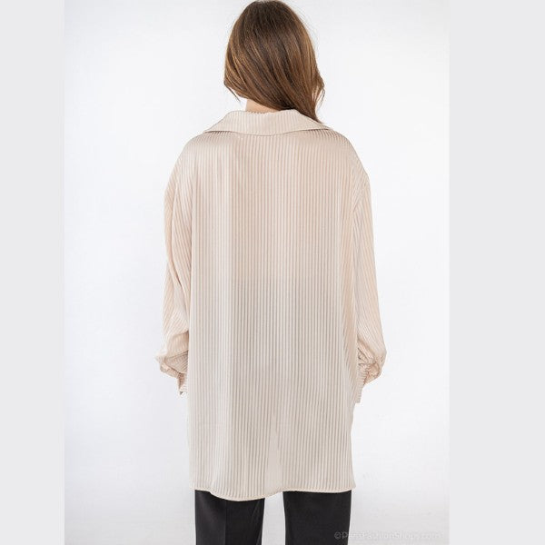 En person med langt, lysebrunt hår står med ansigtet vendt mod kameraet iført en løs, beige J&H Fashion-skjorte med krave og sorte bukser på en almindelig hvid baggrund.