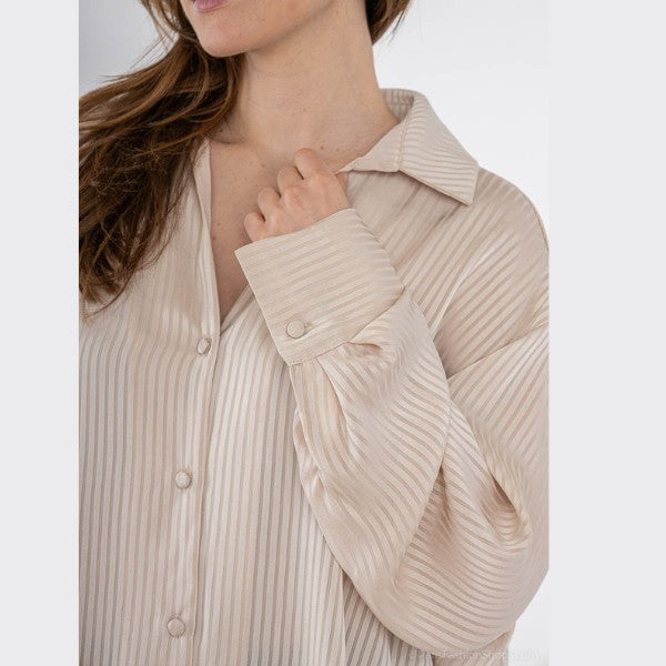En kvinde bærer en løs, beige lodretstribet skjorte fra J&H Fashion. Den silkebløde skjorte har brede manchetter med knapper. Hun holder let i kraven, mens hendes lange brune hår falder ned over den ene skulder, og hendes ansigt er delvist ude af billedet.