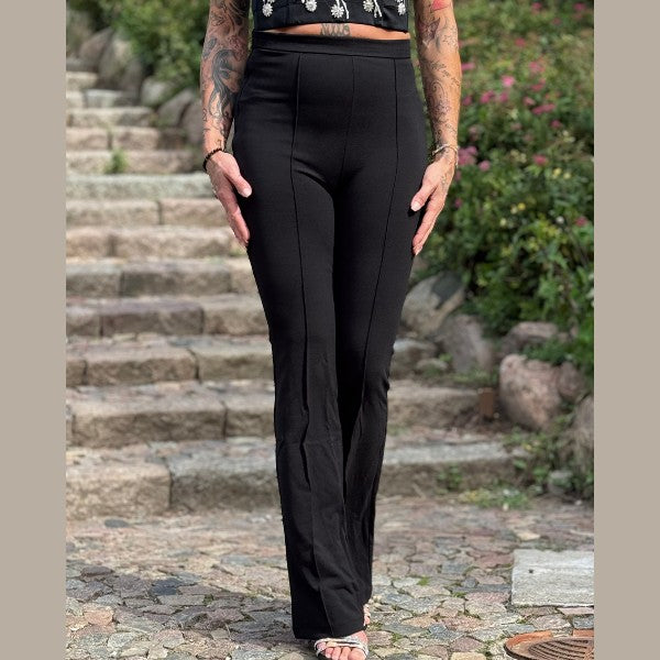 En person står udendørs på en stensti iført MOCHY højtaljet sorte bukser og en sort crop top. Med tatoverede arme og hænder i siden er personen omgivet af grønne buske og blomster. Ansigtet er ikke synligt.