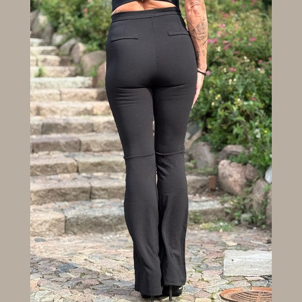 Udendørs på en stensti står en person med ansigtet vendt mod kameraet iført MOCHY sorte bukser med flæser og en sort top. Grønne områder og sten danner baggrund, og man kan se deres tatoveringer på armene.