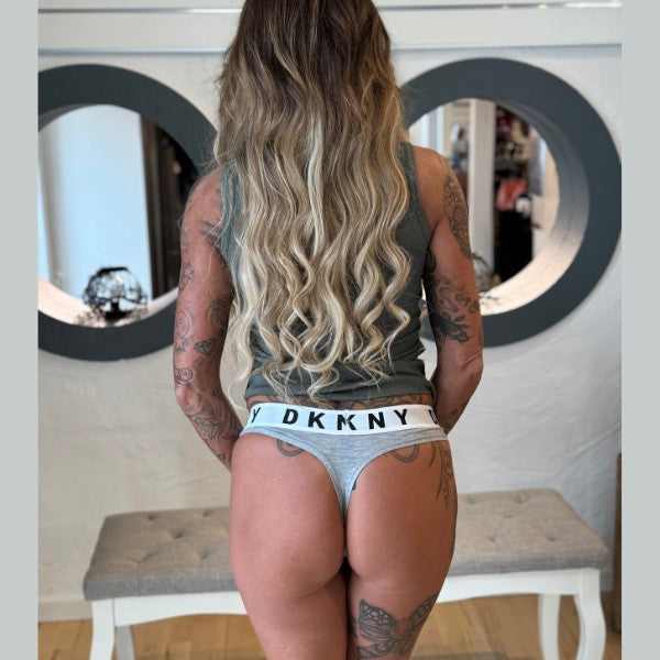 En kvinde med langt, bølget blond hår vender ansigtet væk fra kameraet, iført en grå ærmeløs top og DKNY Cozy Boyfriend Thong-undertøj. Hendes tatoverede arme og ryg giver et edgy touch, mens hun står ved siden af en bænk og to runde spejle.