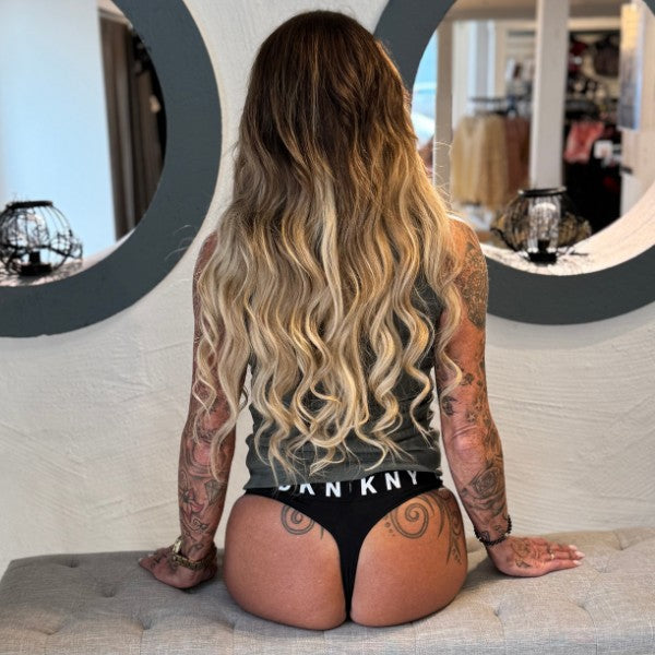 En kvinde med langt, bølget ombréblondt hår sidder på en bænk iført en grå tanktop og sorte DKNY Boyfriend Thong, hendes tatoverede arme og ryg er synlige. Det sporty outfit står i kontrast til det minimalistiske design og de lysende lys.