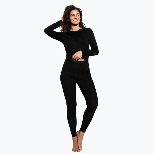 En mørkhåret kvinde smiler barfodet mod en almindelig lys baggrund iført en tætsiddende langærmet sort top og Panos Emporio-leggings. Hun poserer med den ene hånd på hoften og den anden på hovedet.