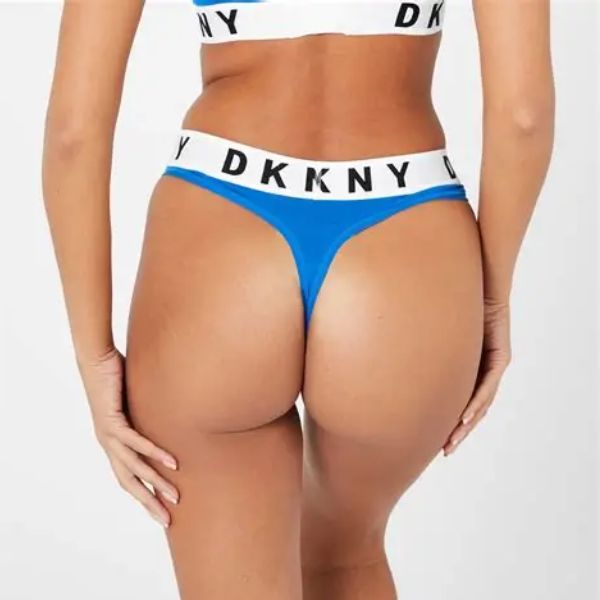 En kvinde ses fra taljen og ned, med ryggen til, i en blå stringtrusse og matchende hvid DKNY-bralette. Hendes venstre hånd hviler på hoften og fremhæver den sporty linning med et fedt DKNY-branding på en almindelig hvid baggrund.