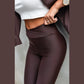 En person bærer Ivon-leggings med høj talje og elastisk pasform i chokoladebrun, en hvid kort tanktop og en brun jakke, der er trukket over skuldrene. Et guldur og en vielsesring pryder håndleddet. Baggrunden er almindelig og neutral.