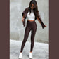 En kvinde står op ad en grå væg iført Ivon høj talje leggings, en kort hvid top, en brun jakke over skuldrene, hvide sneakers og solbriller. Hun poserer med den ene fod fremme og kigger ned med løsthængende hår, der indrammer hendes ansigt.