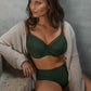 En kvinde med bølget brunt hår står model til en mørk grøn trusse og matchende bh fra Fantasie. Hun har en beige cardigan over skuldrene, mens hun sidder på en betontrappe og kigger til siden mod en neutral grå baggrund.